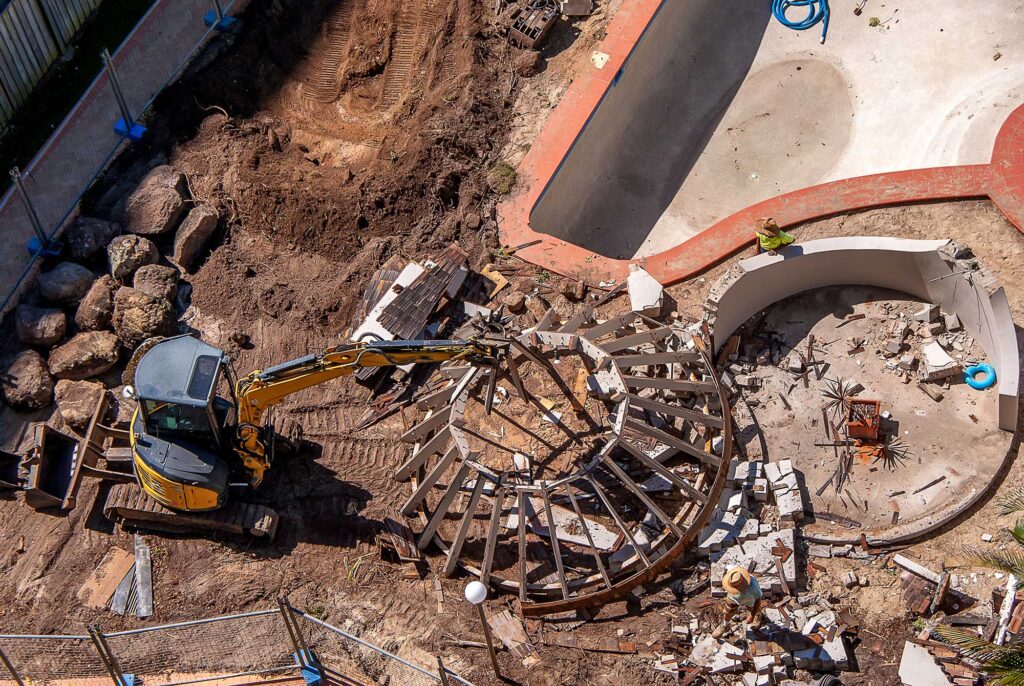 An aerial view of an excavator destroying an old swimming pool.
